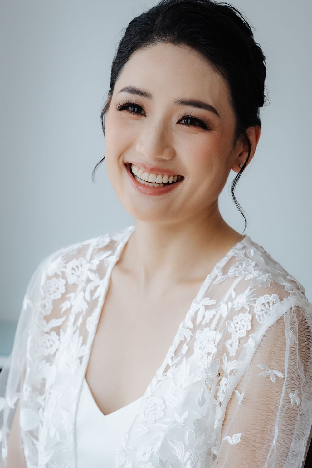 Smiling Asian bride in white lace robe with elegant updo hairstyle and natural makeup on her wedding day.