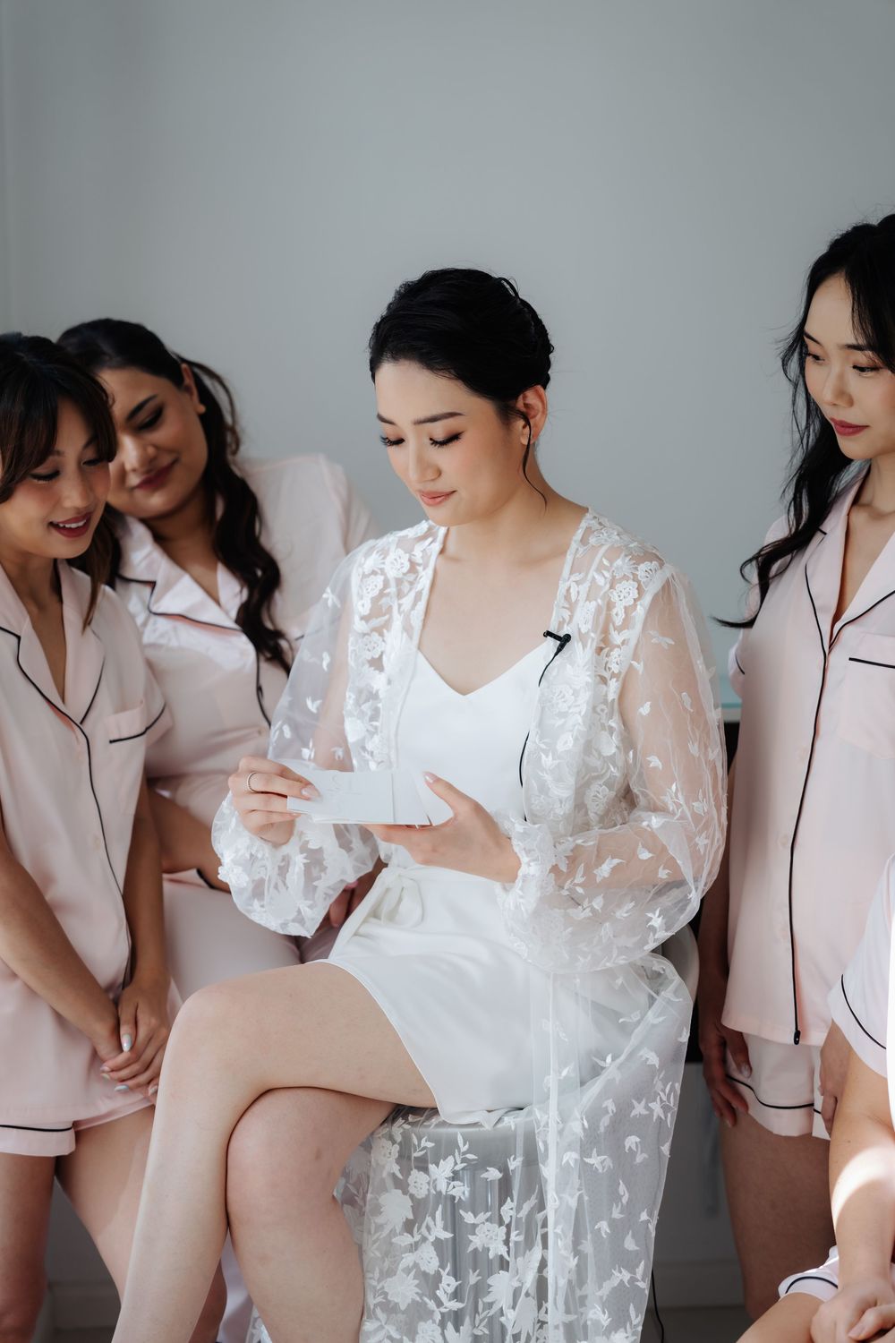 Bride in white lace robe reads a note surrounded by bridesmaids in pink satin pajamas during wedding morning prep.