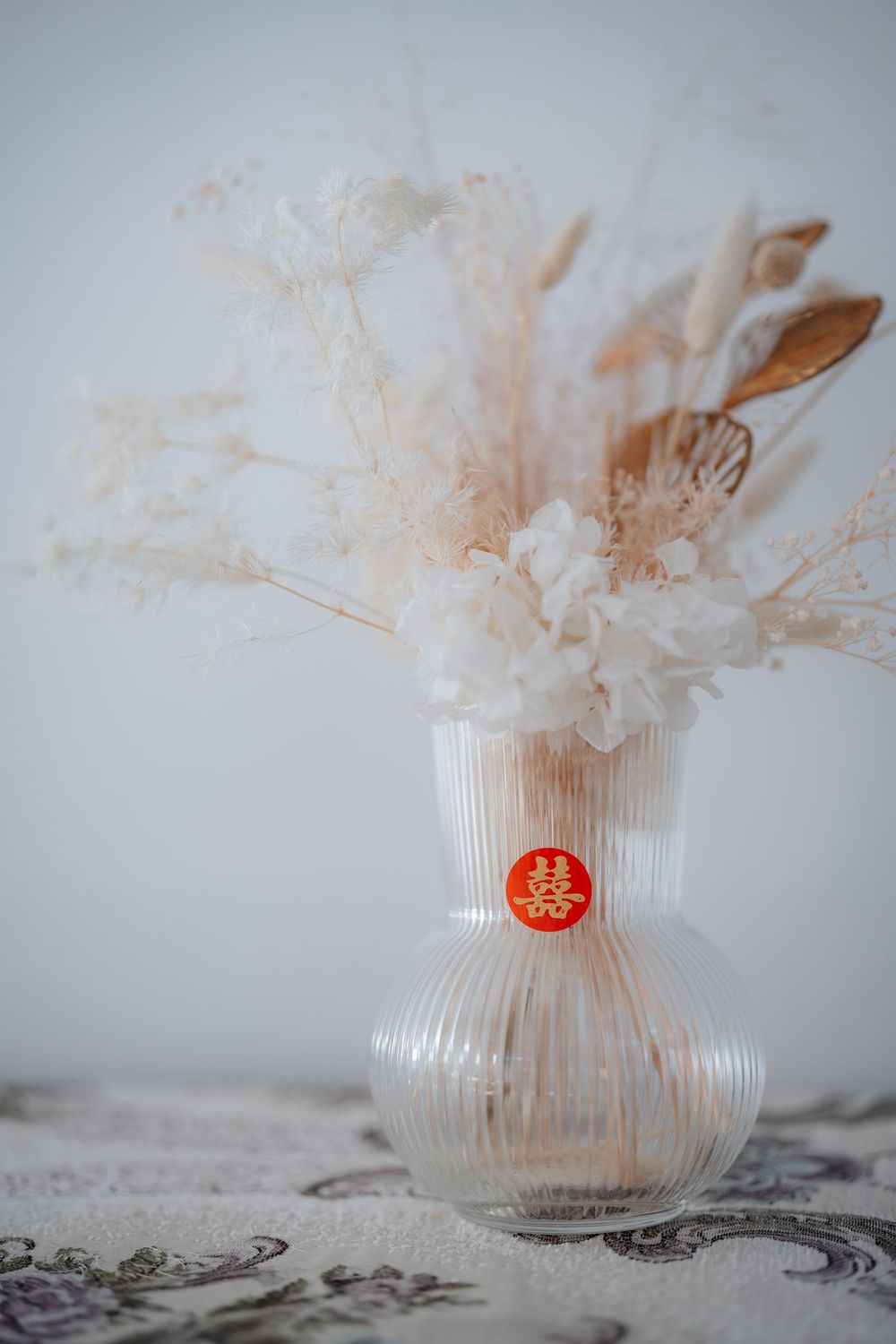 Elegant dried floral arrangement in ribbed glass vase with Chinese double happiness symbol, featuring white hydrangeas and pampas grass.