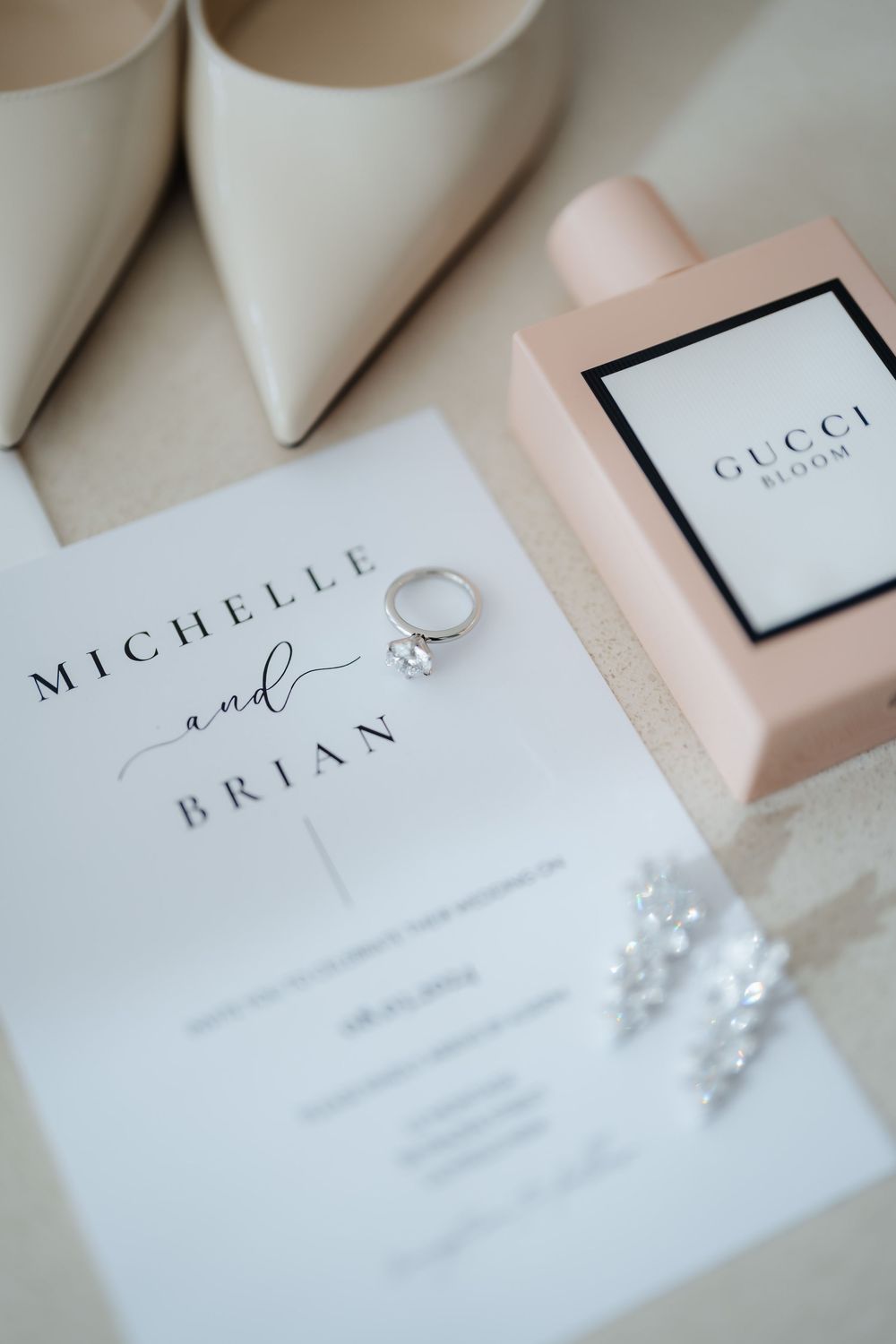 Wedding flat lay featuring Michelle and Brian invitation, diamond ring, pearl earrings, Gucci Bloom perfume, and ivory heels.