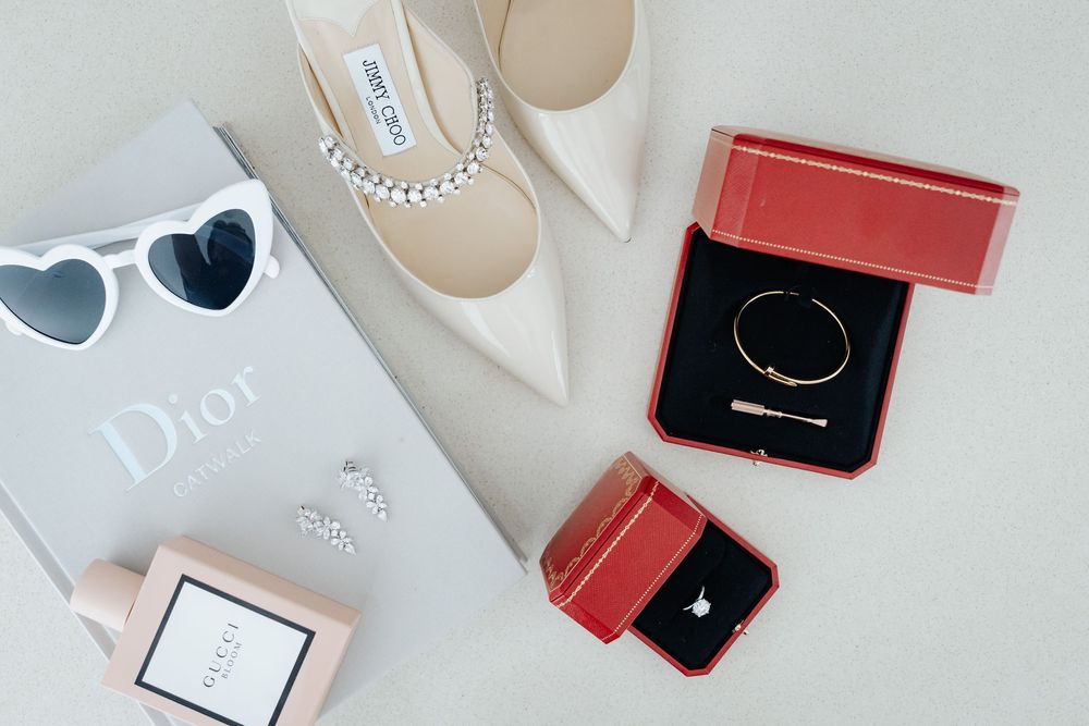 Luxury bridal accessories flatlay featuring Dior bag, Jimmy Choo heels, Gucci perfume, heart sunglasses, and Cartier jewelry boxes.