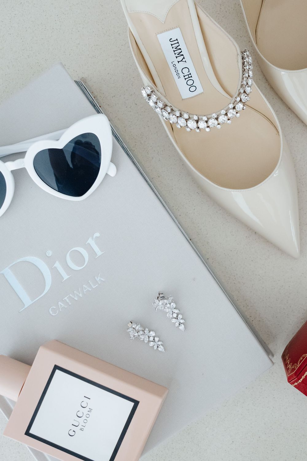 Luxury flat lay featuring Jimmy Choo heels, Dior Catwalk book, Gucci perfume, heart sunglasses, and crystal earrings.