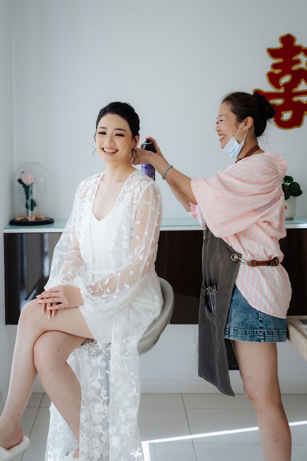 A bride in a white lace robe smiles while a makeup artist in a pink top and mask styles her hair indoors.
