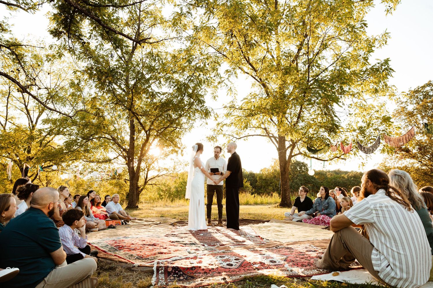 Couple shares first dance at golden hour outdoor wedding ceremony with guests seated on rugs under trees.