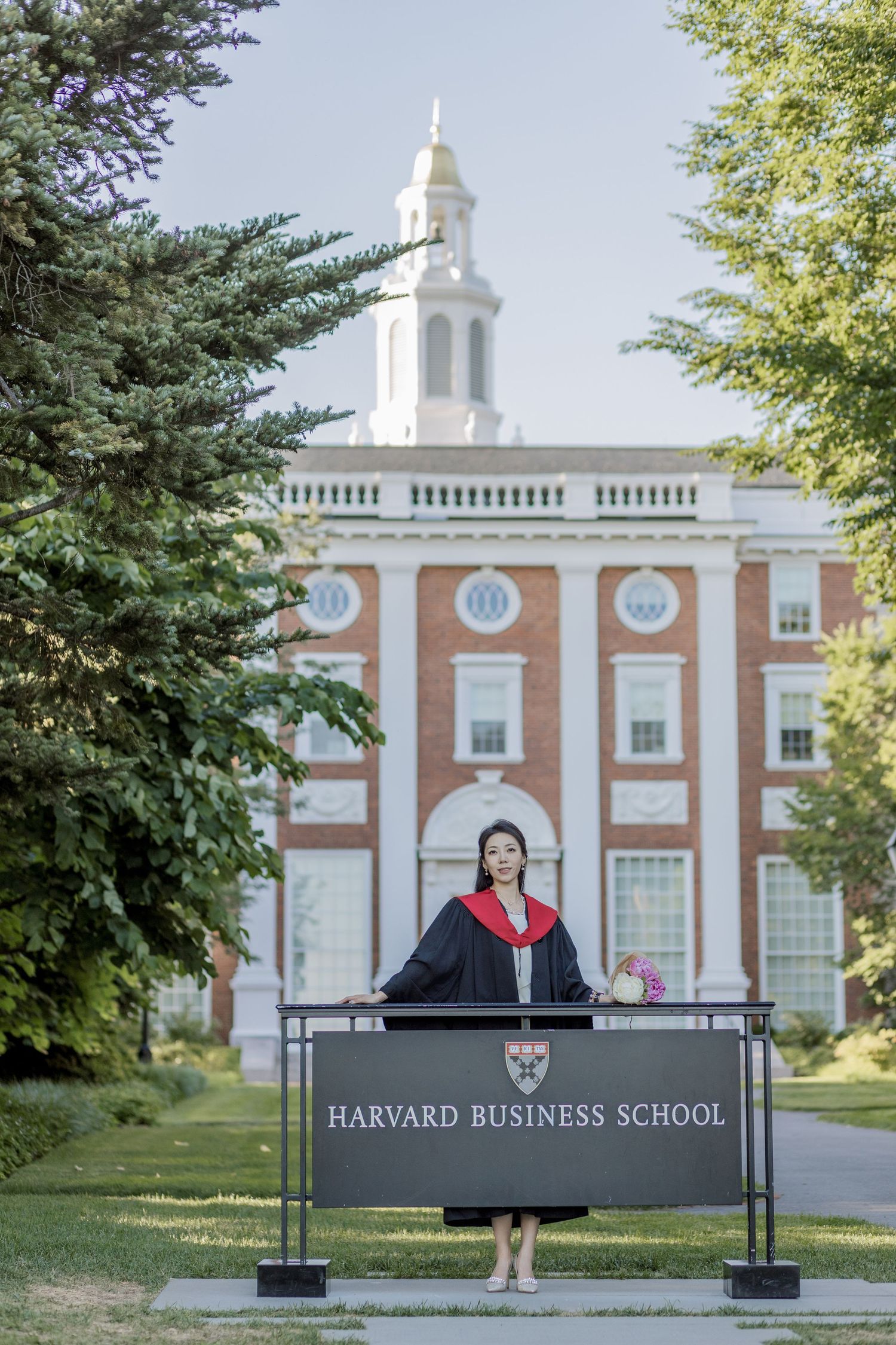Harvard Graduate in academic regalia stands at Harvard Business School podium with iconic brick building backdrop.