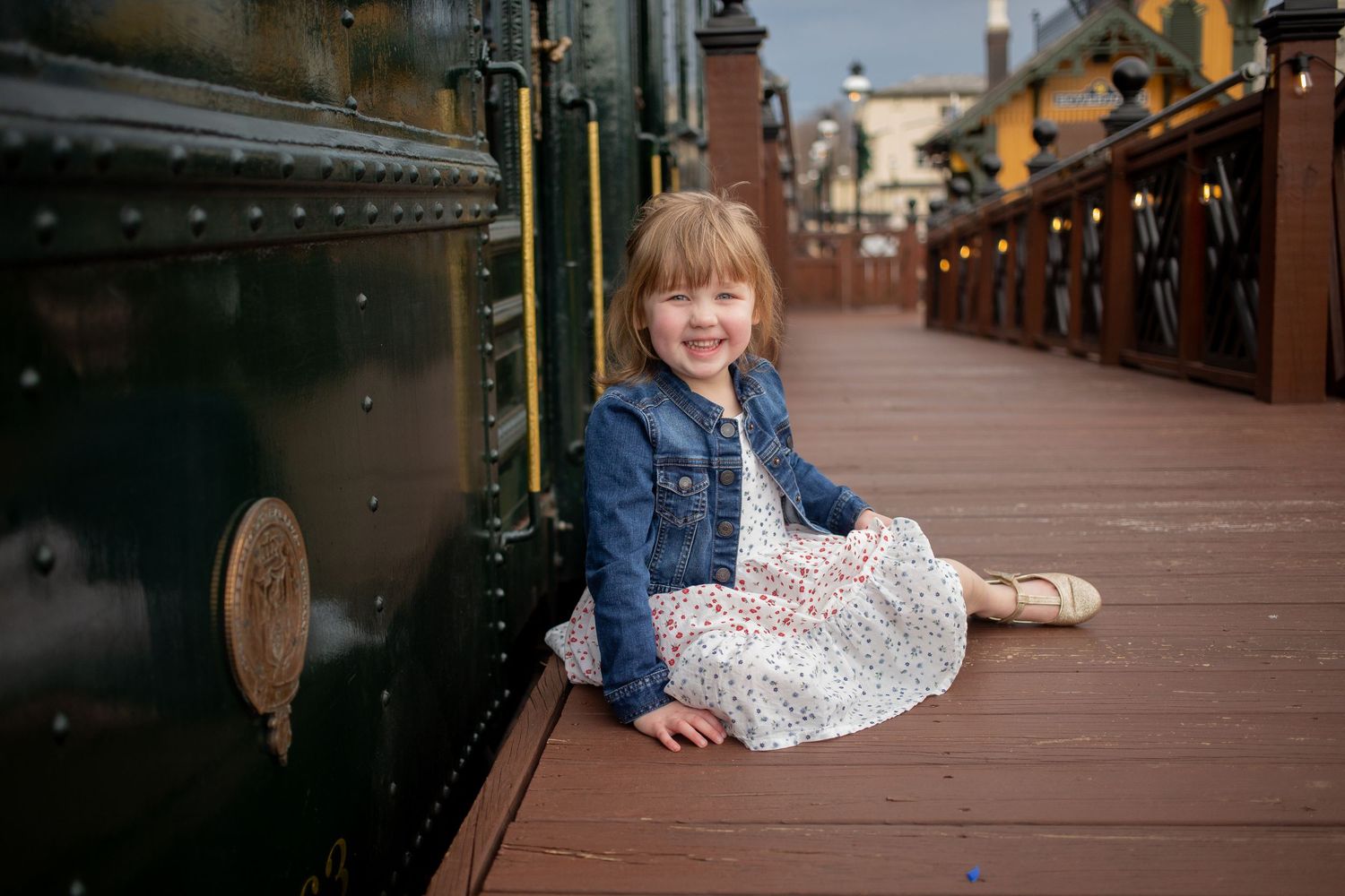 Young girl in denim jacket and floral dress sitting beside a vintage train, smiling joyfully on a wooden platform.