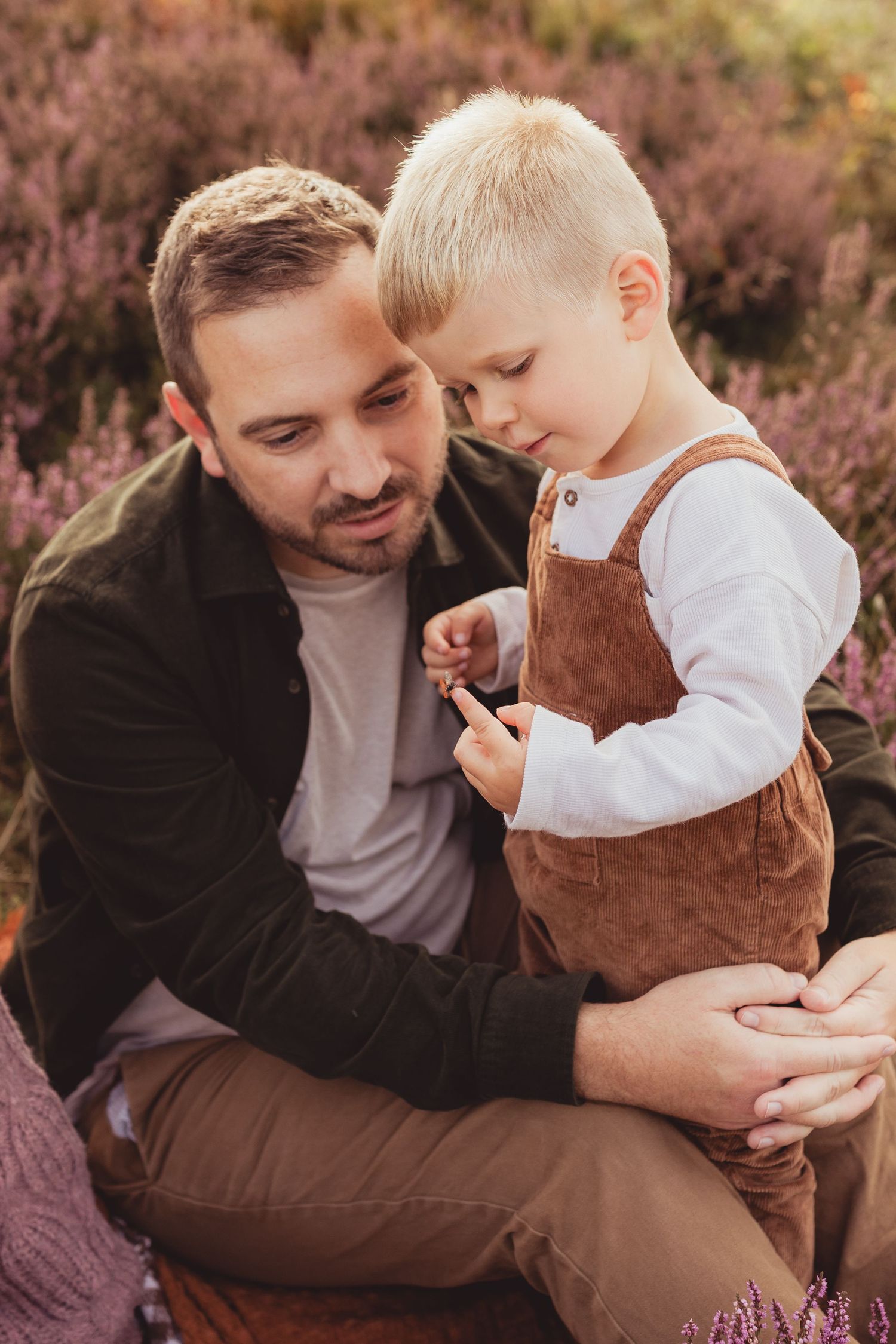 A father sits outdoors in a heather field, lovingly holding his young blonde son in brown overalls.