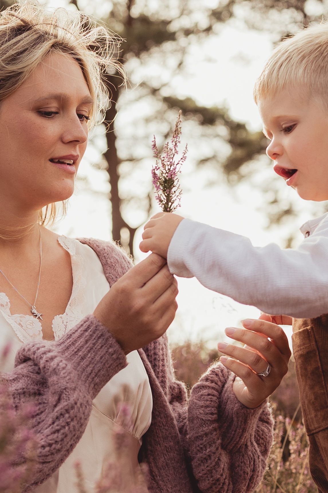 A mother and toddler share a tender moment outdoors, examining a small pink wildflower together in soft natural light.