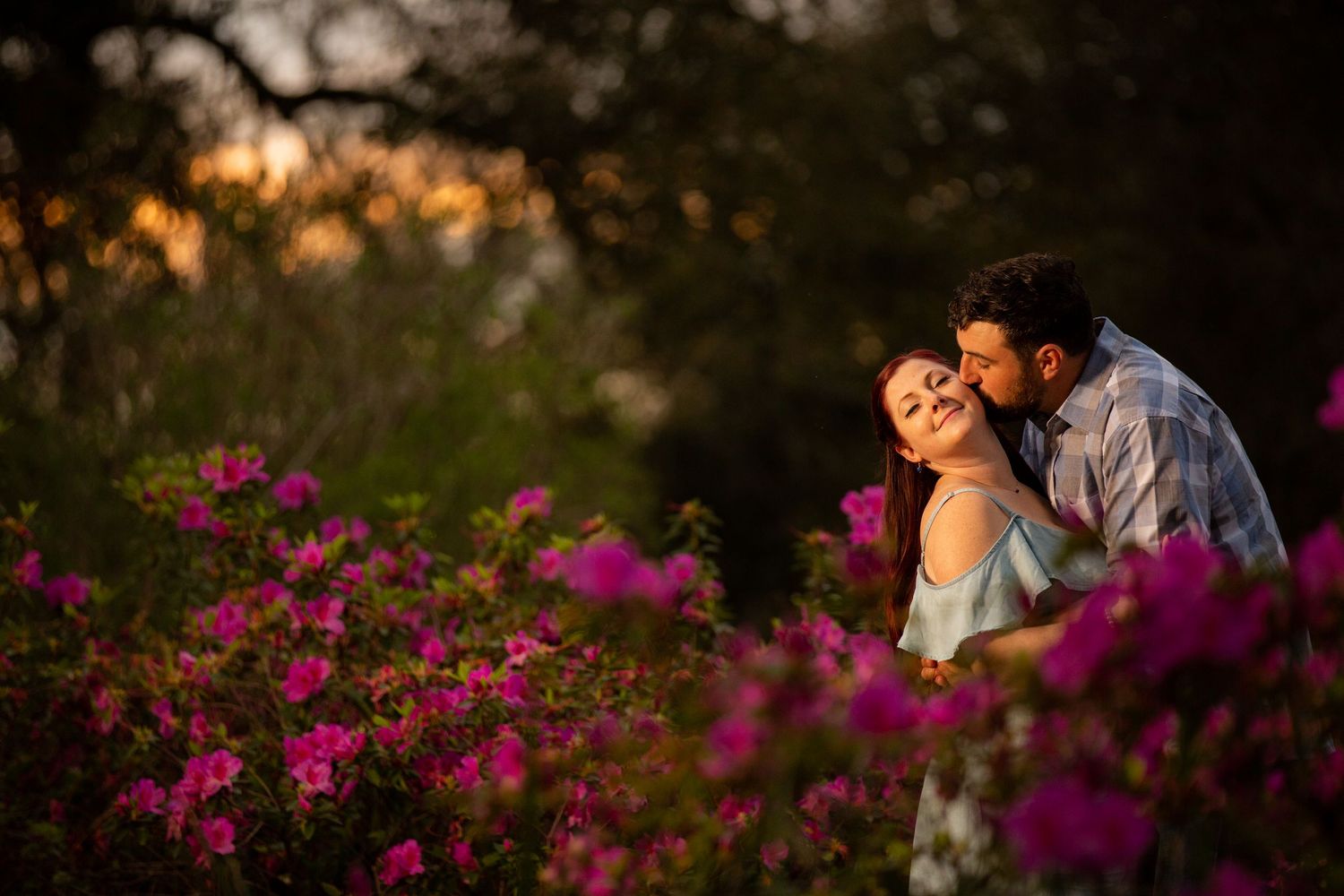 Couple sharing a romantic kiss among vibrant pink flowers at golden hour in a lush garden setting.