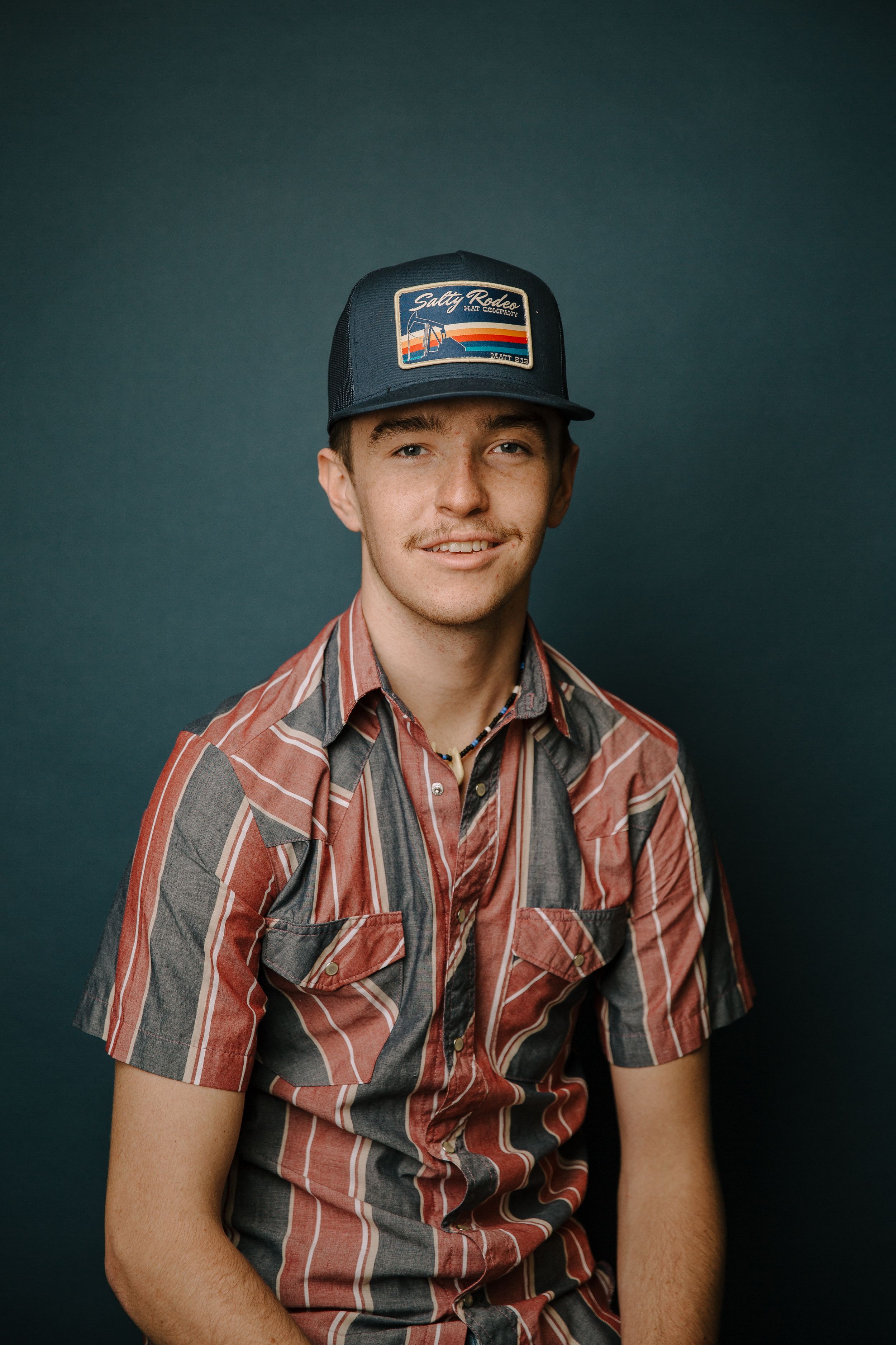 Young man smiling in striped shirt and navy trucker hat poses against teal background.