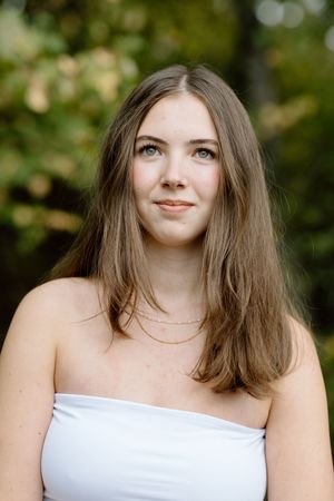 Young woman with long brown hair and blue eyes wearing a white strapless top and gold necklace outdoors.