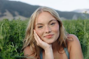 Young blonde woman with freckles smiling outdoors, resting chin on hand among lush green plants with mountains behind.