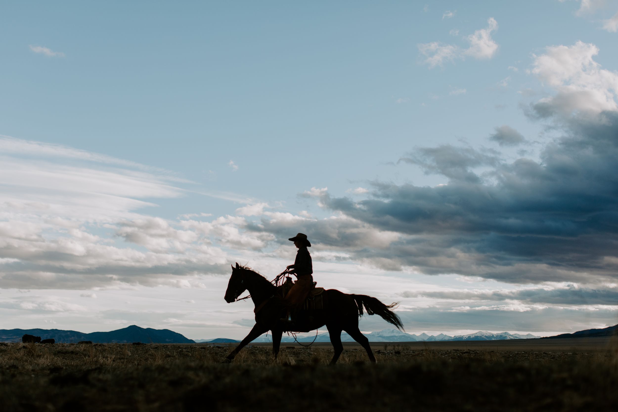 Silhouette of a cowboy riding a horse across an open field at dusk with dramatic cloudy sky and mountains.