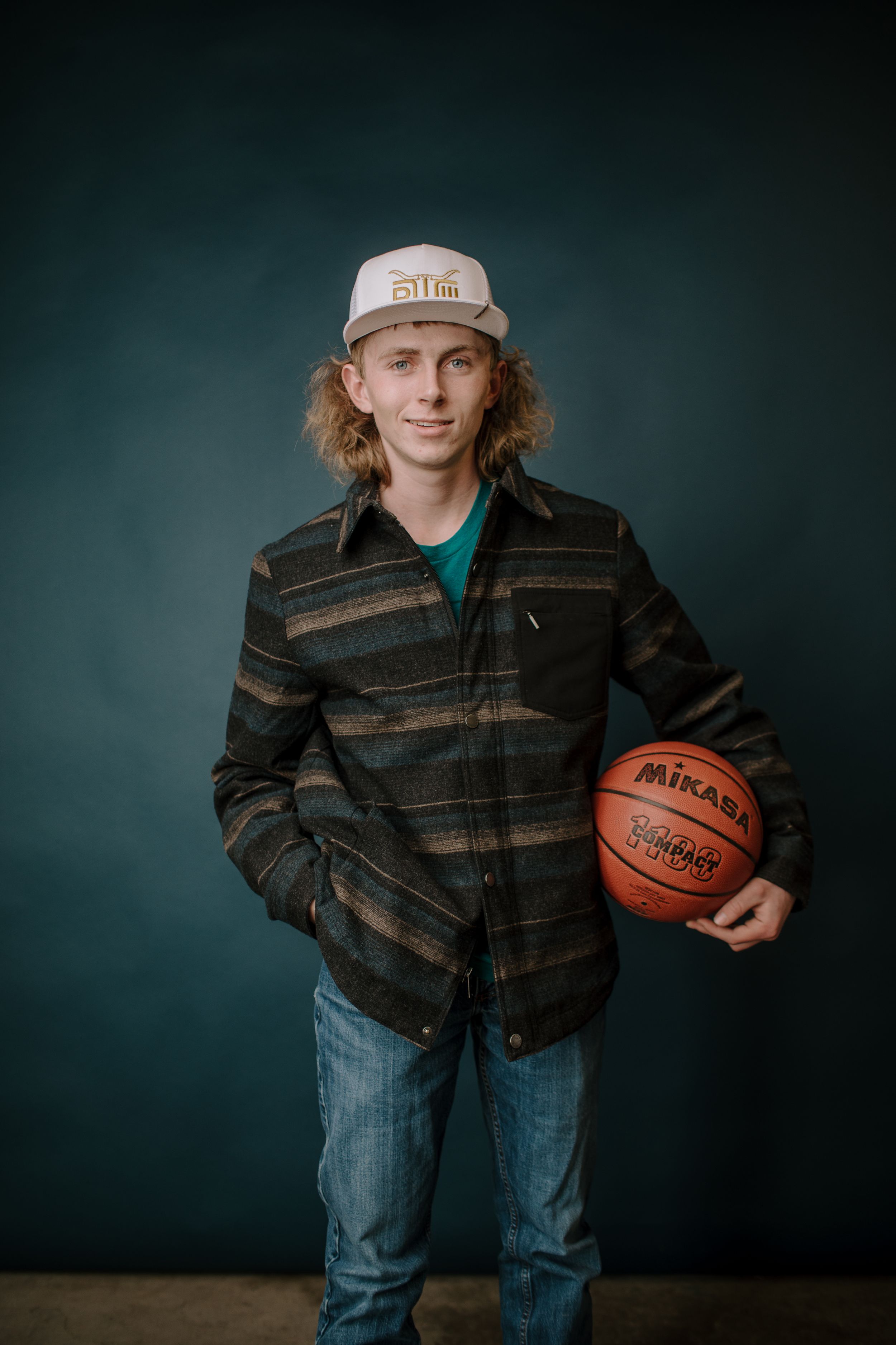 Young man with curly hair holding a Mikasa basketball, wearing a flannel shirt and trucker hat, posing against a dark teal backdrop.