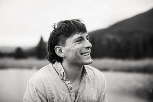Black and white portrait of a young smiling man outdoors near a lake with mountains in the background.