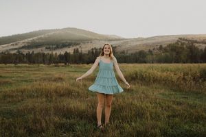 Young woman in teal dress twirling in green meadow with rolling hills and pine trees at golden hour.