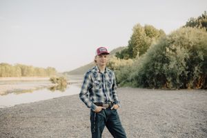 Young man in plaid shirt and red trucker cap standing on rocky riverbank with trees in background.
