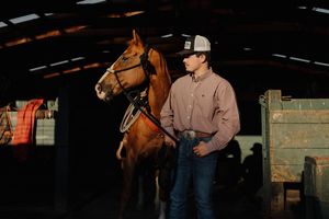 Young cowboy in a pink shirt and cap stands beside a chestnut horse inside a rustic barn at golden hour.