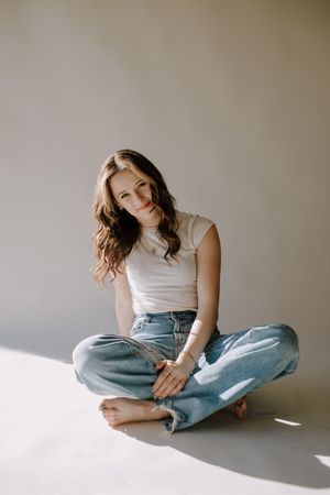 Young woman with wavy brown hair sits cross-legged on floor wearing white tee and blue jeans, smiling softly.