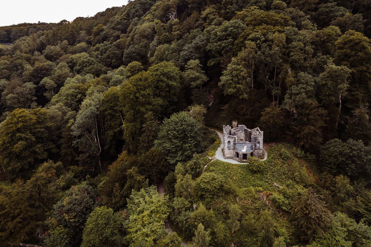 Couple poses in wedding attire atop at the Claife Viewing station surrounded by lush green trees and foliage.