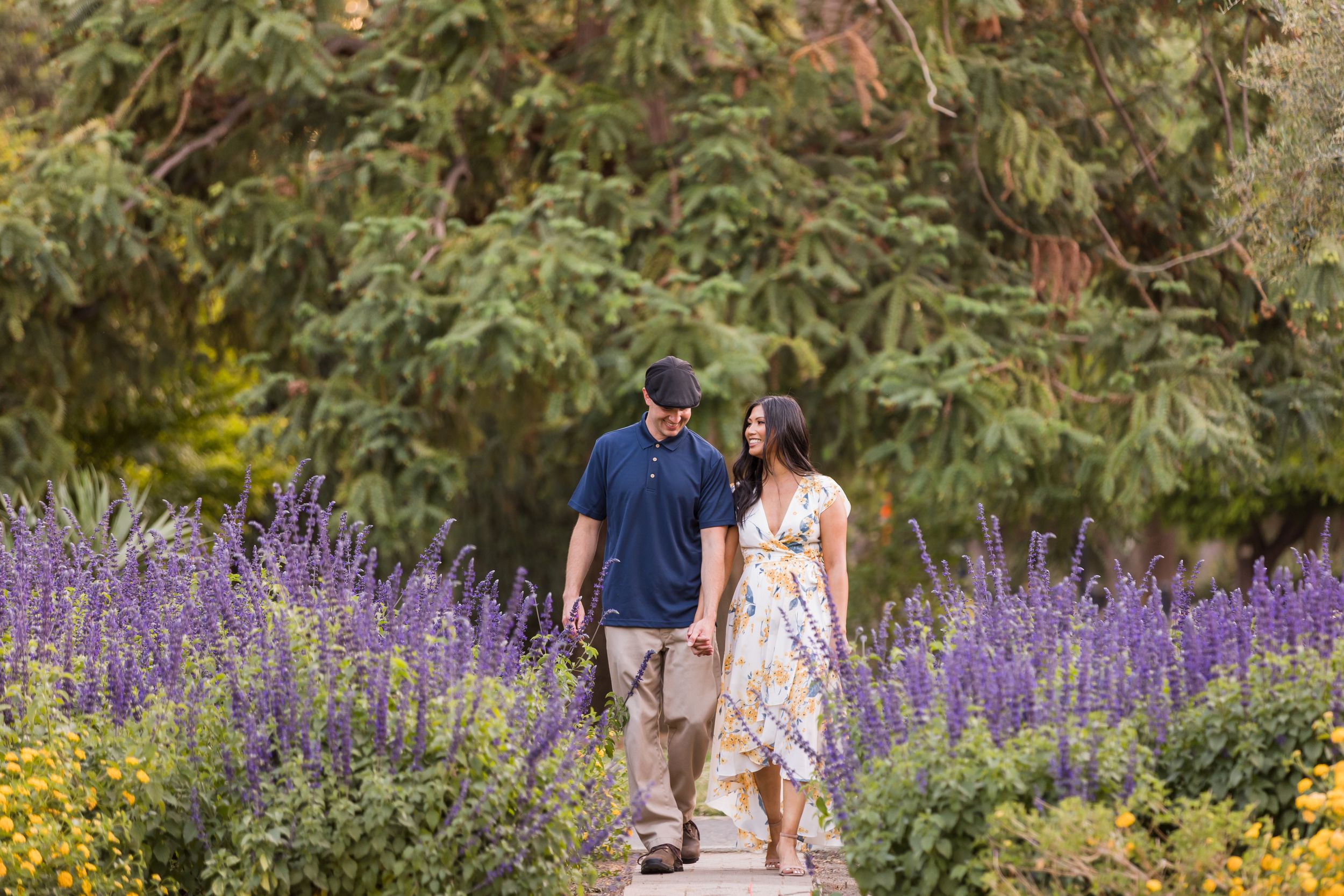 Couple walking hand-in-hand on a garden path surrounded by purple lavender and yellow flowers with lush green trees.