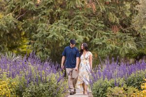Couple walking hand-in-hand on a garden path surrounded by purple lavender and yellow flowers with lush green trees.