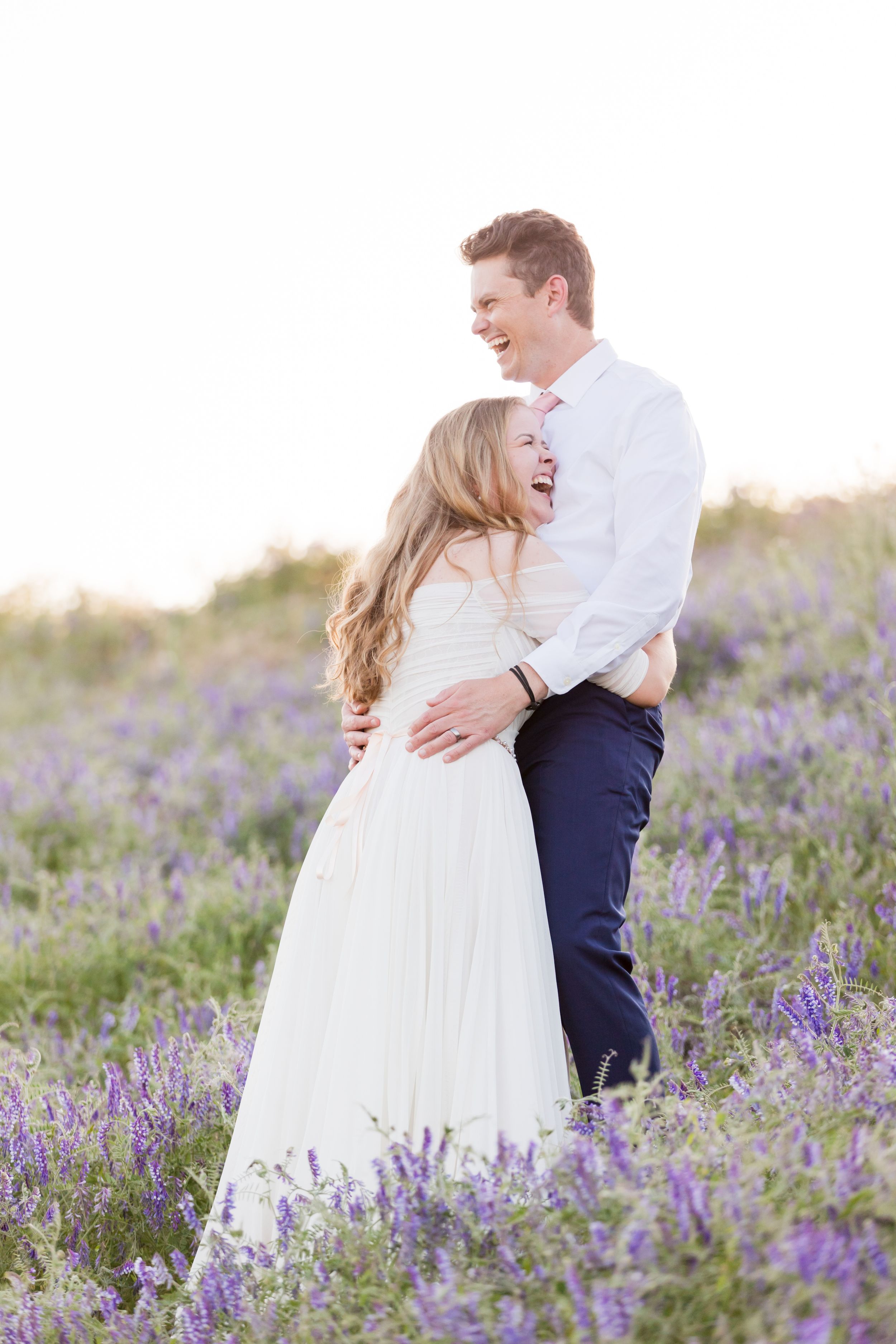 A joyful couple embraces and laughs in a blooming lavender field, bride in white gown and groom in white shirt.