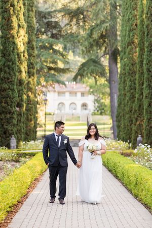Bride and groom walk hand-in-hand along a tree-lined garden path toward an elegant estate on their wedding day.