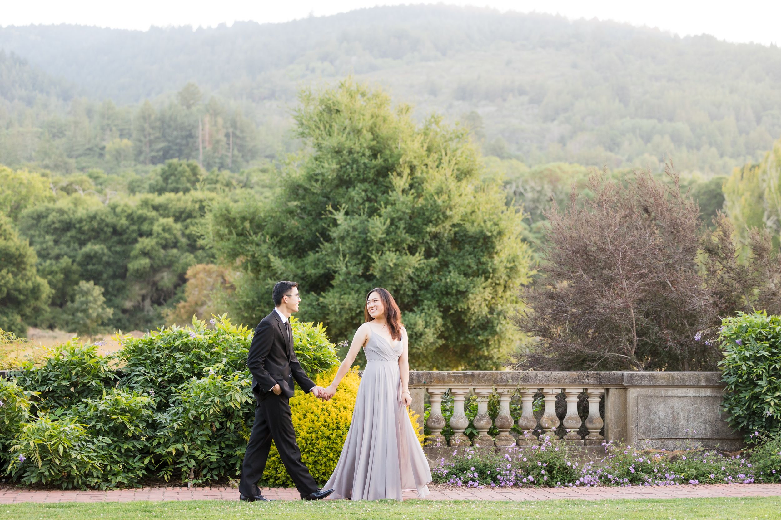 Couple walks hand-in-hand in lush garden, woman in flowing gray gown, man in black suit, green hills backdrop.