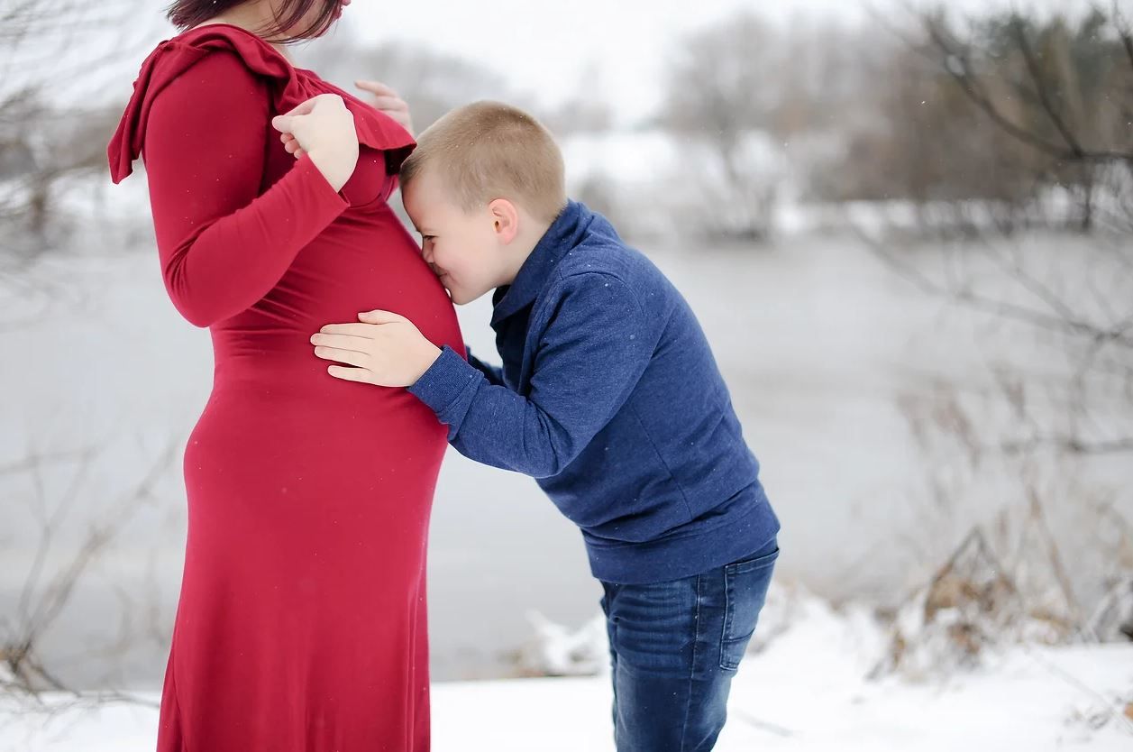 A young child in blue embraces a pregnant belly in a snowy winter setting.