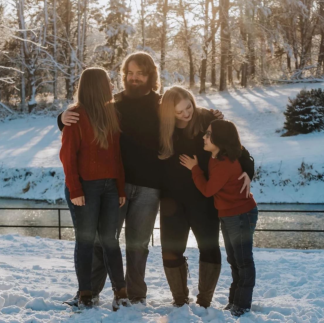 Four people embrace while standing in fresh snow with bare trees and winter sunlight behind them.