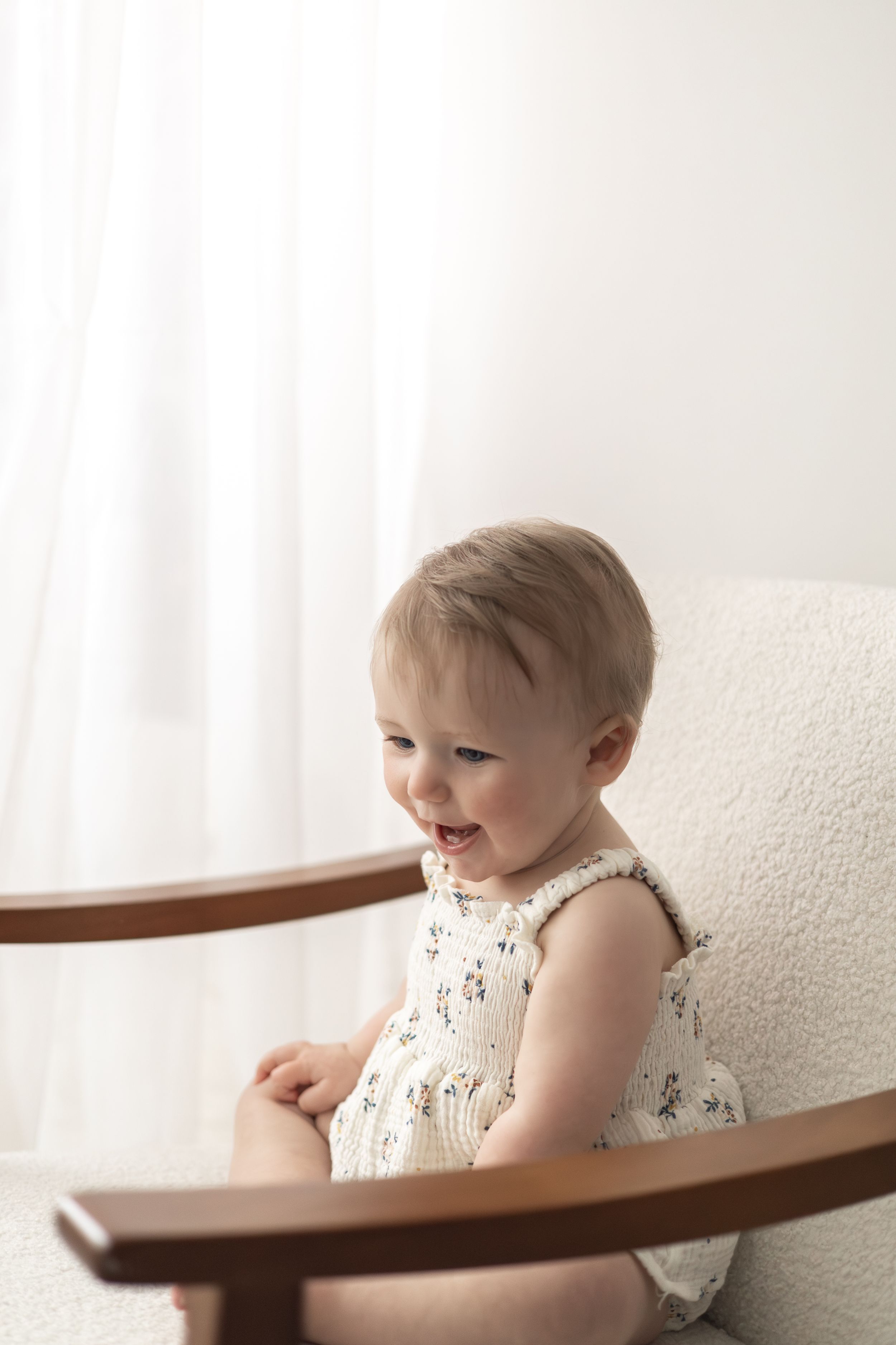 Smiling baby girl in floral dress sitting in a wooden chair against a white background Nottingham first birthday photography baby milestone