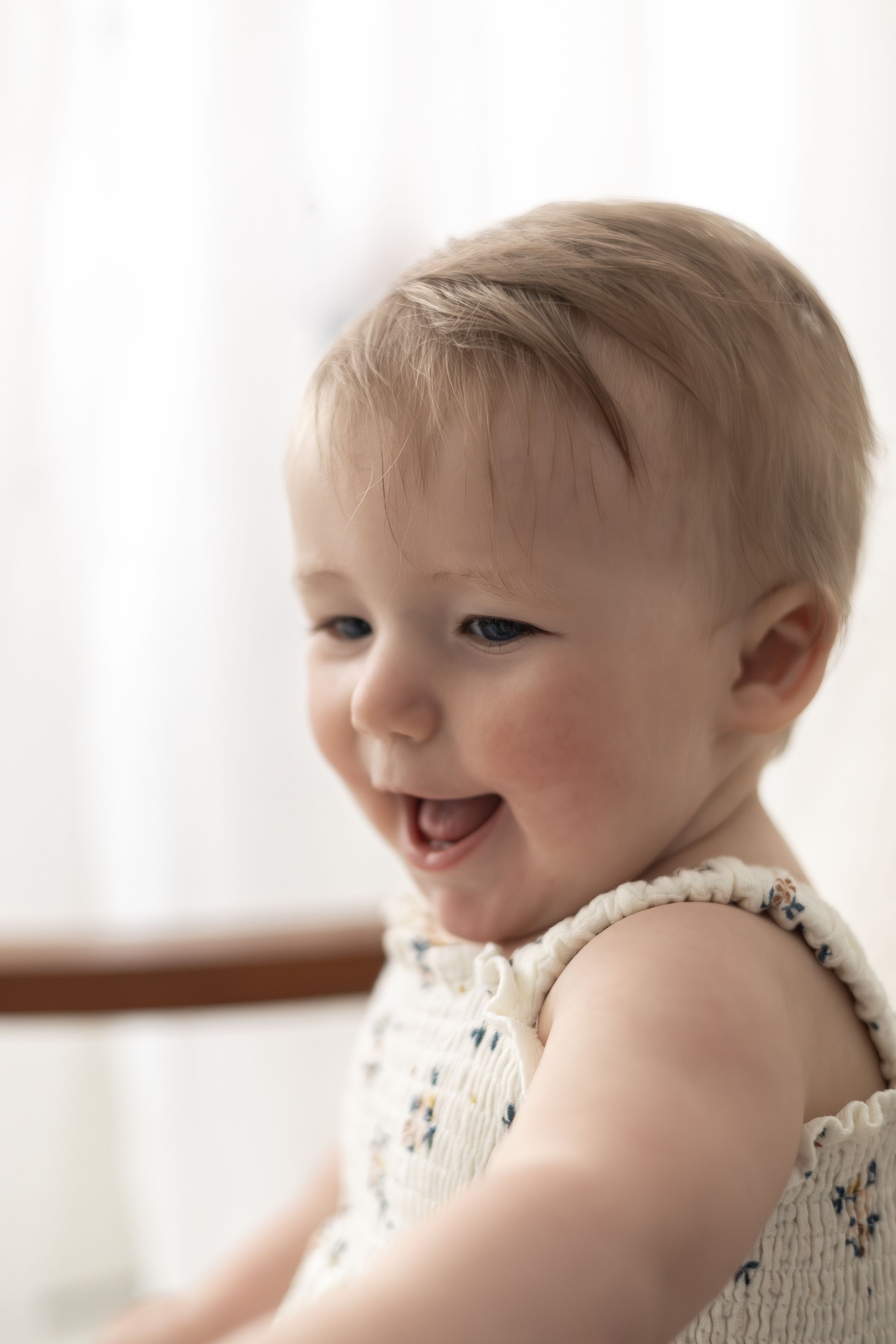 A happy laughing baby girl in a floral romper sits in soft natural light Nottingham baby milestone first birthday photography