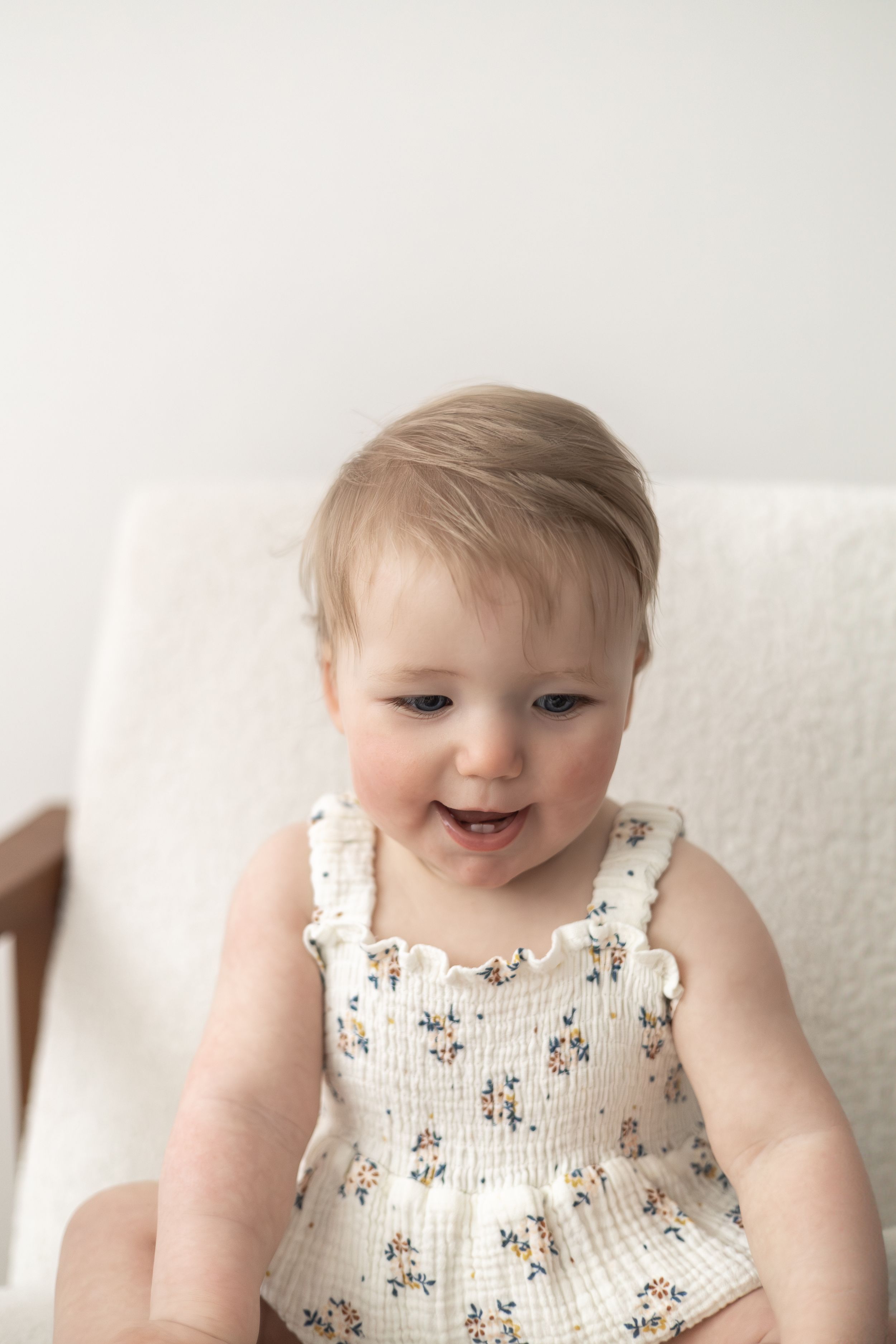 Smiling baby girl in a white floral sundress sitting in a chair against a light background first birthday photography Nottingham