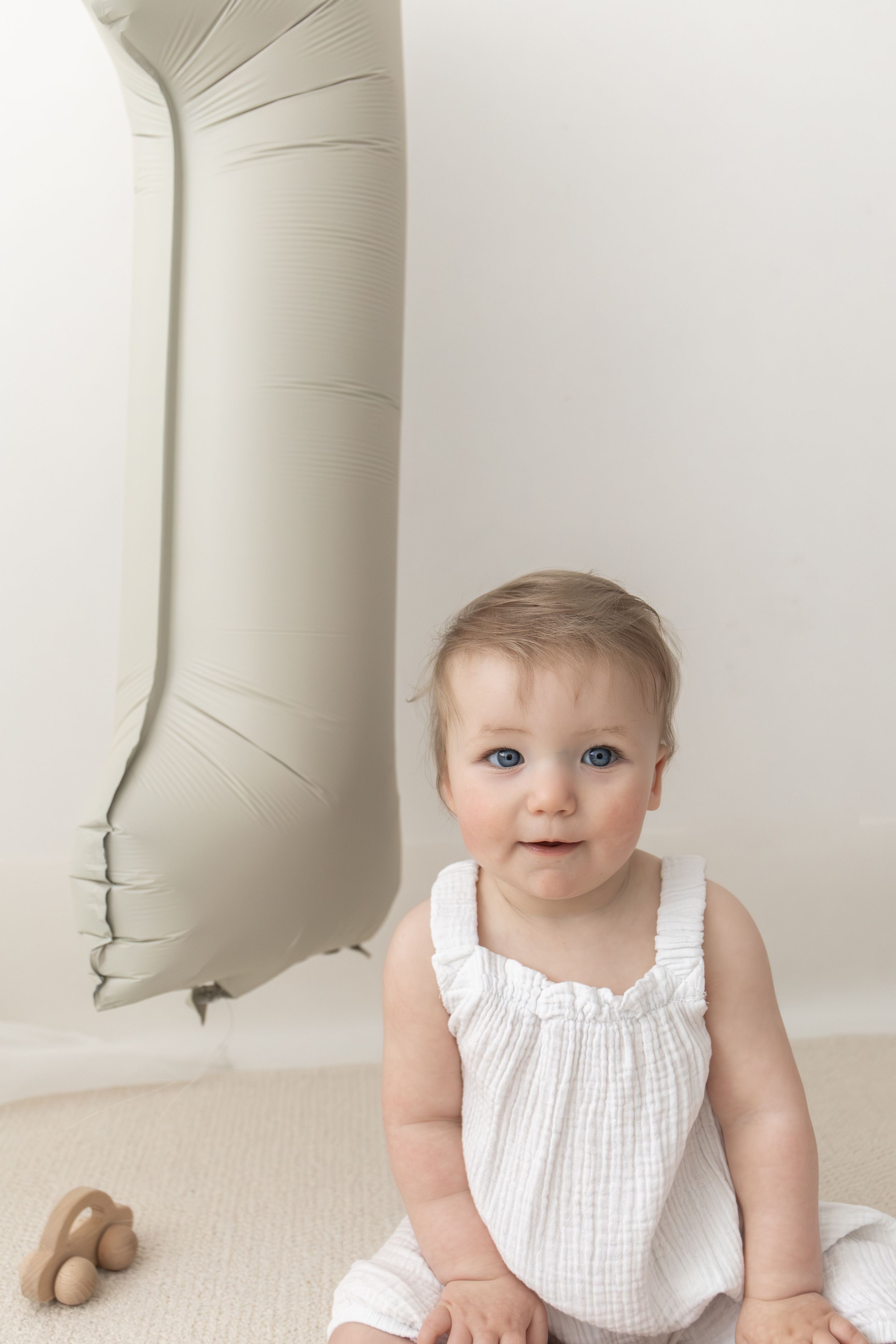 Baby in white dress sits beside a number 1 balloon, celebrating their first birthday milestone Nottingham first birthday photography