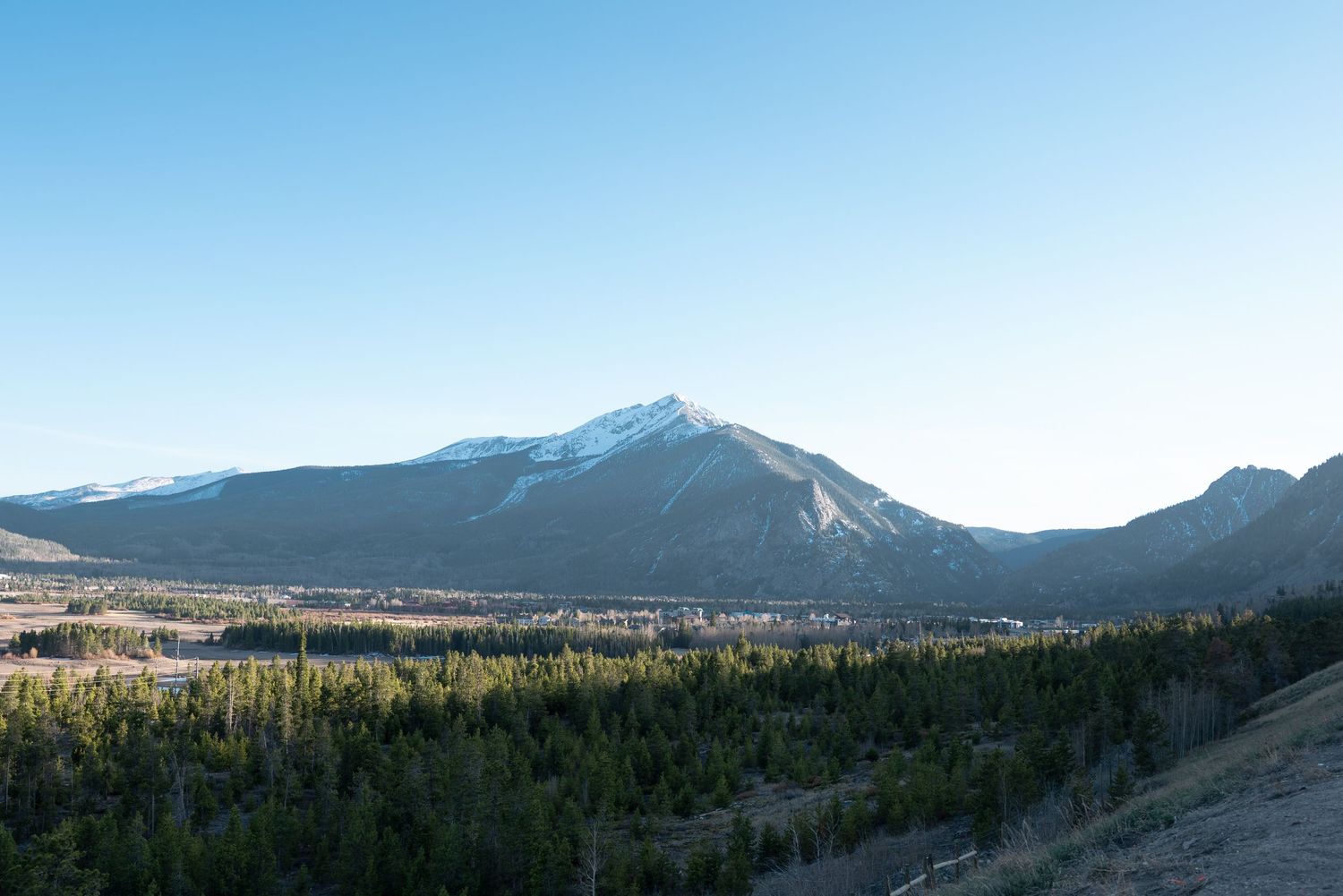Snow-capped mountain peak rises above dense evergreen forest under clear blue sky in scenic Rocky Mountain landscape.