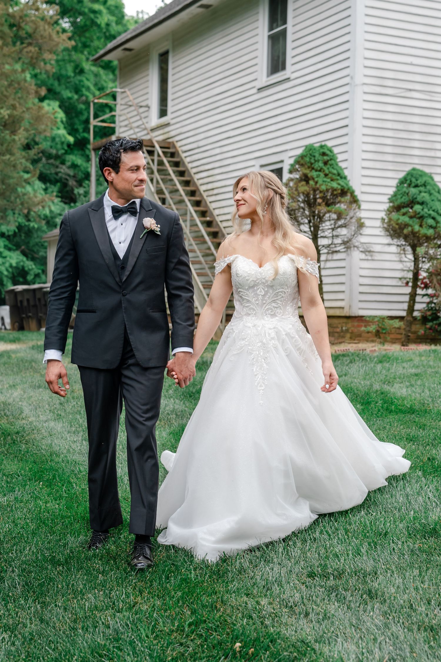 A bride and groom walk hand in hand on green grass, smiling at each other, wearing a white gown and black tuxedo.