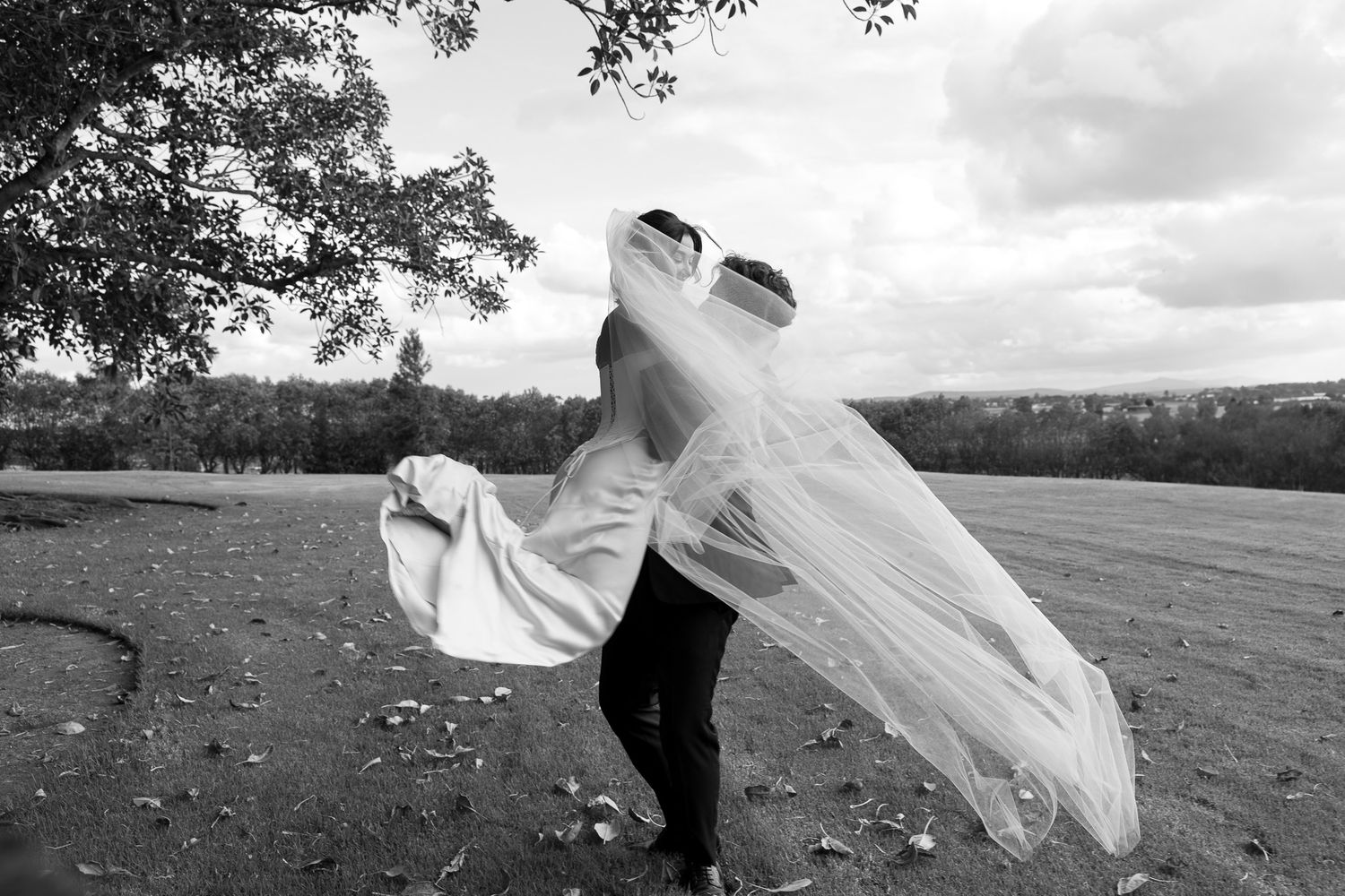Black and white photo of a bride and groom embracing outdoors, her veil billowing in the wind on a cloudy day.