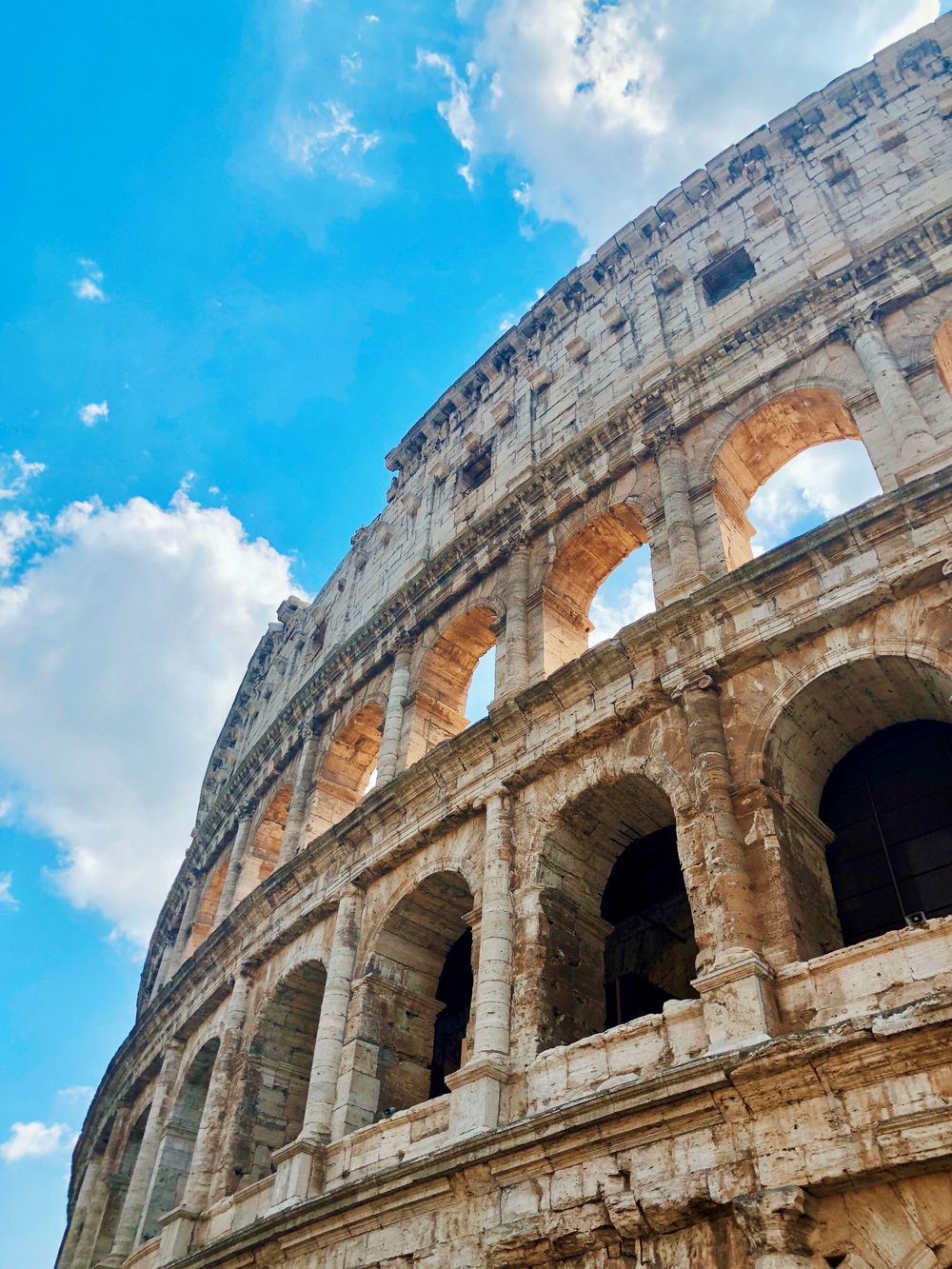 Low-angle view of the ancient Roman Colosseum ruins with arched facades against a bright blue cloudy sky.