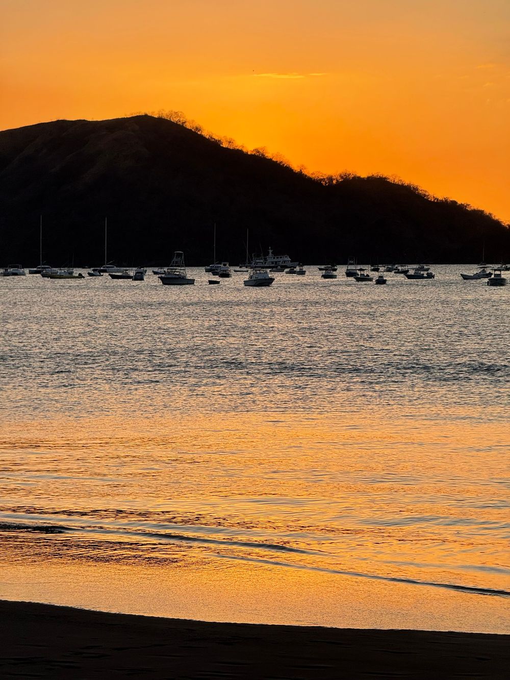 Golden sunset over a tranquil bay with silhouetted boats anchored near a dark hillside, reflecting warm orange hues on calm water.