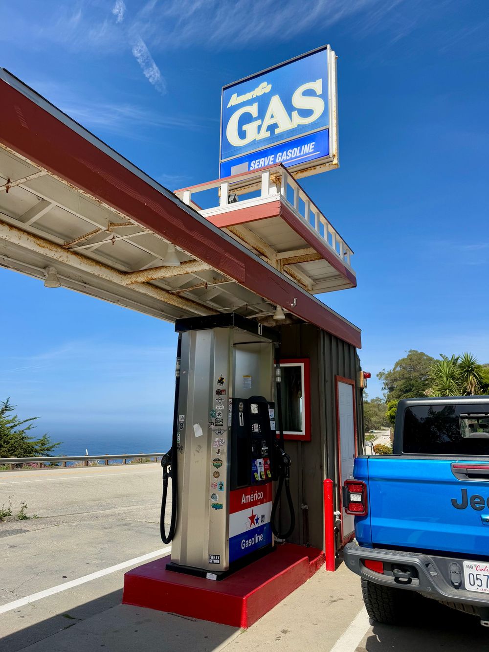 A retro gas station with a blue GAS sign, fuel pumps, and a blue Jeep parked under a canopy with ocean views.