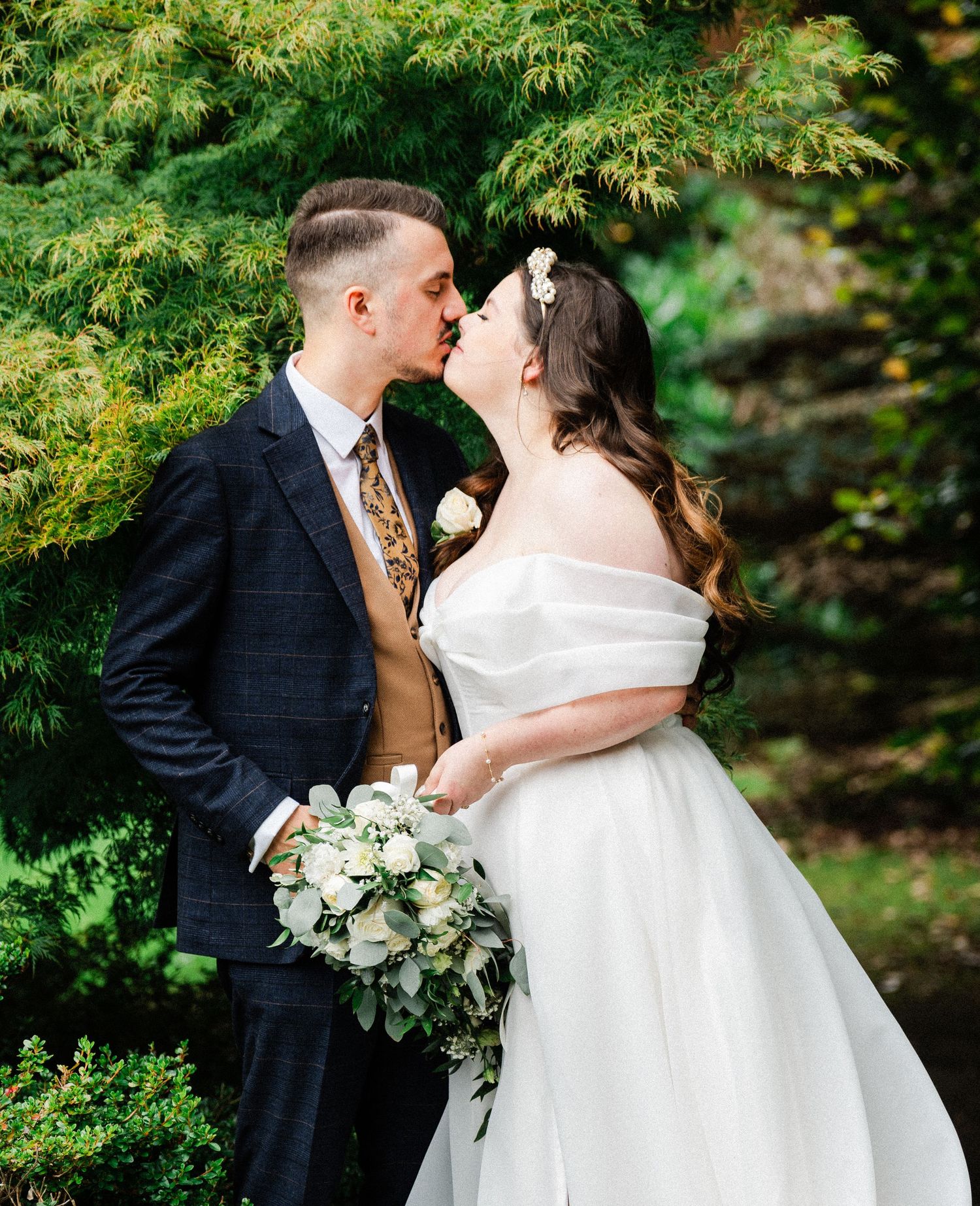 A bride and groom share a kiss outdoors, she holds a white bouquet, he wears a navy suit with gold tie.