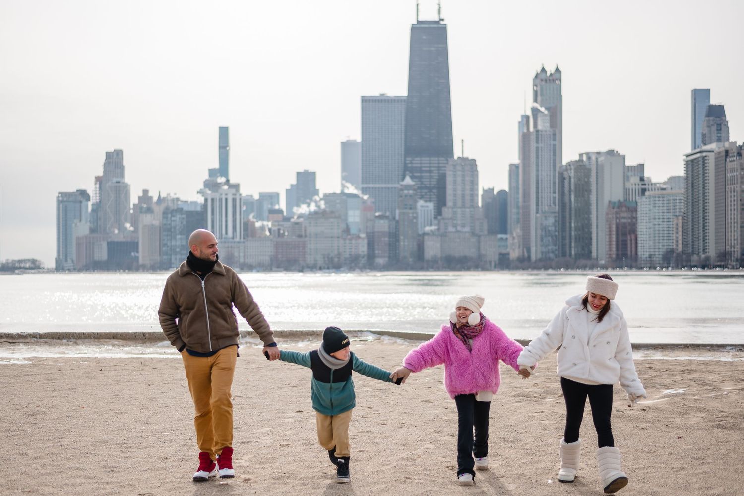 Family of four holding hands walking on a beach with Chicago city skyline in the background on a winter day.