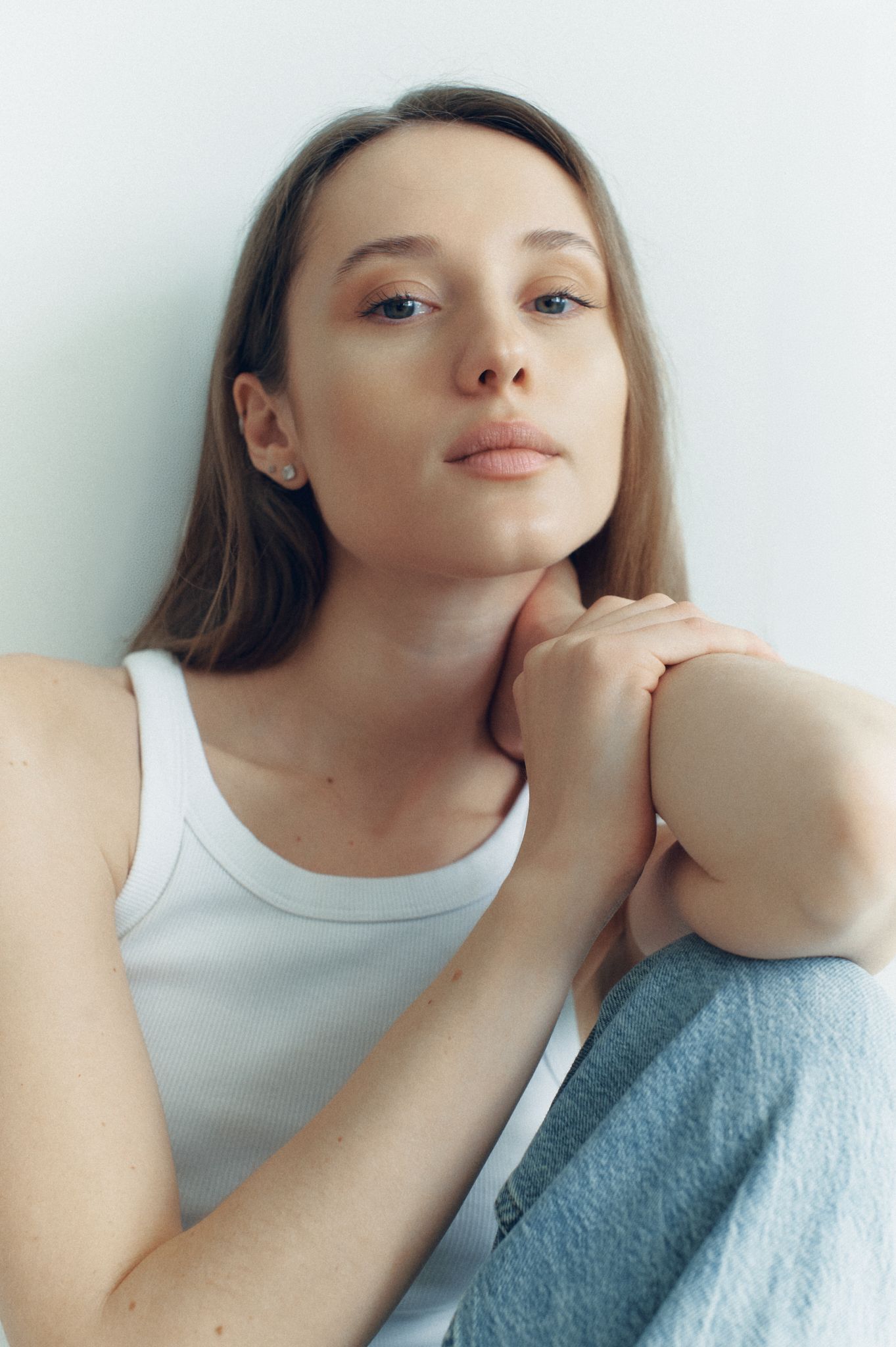 Young woman in white tank top and jeans sitting against a light wall, hands clasped near chin, looking at camera.