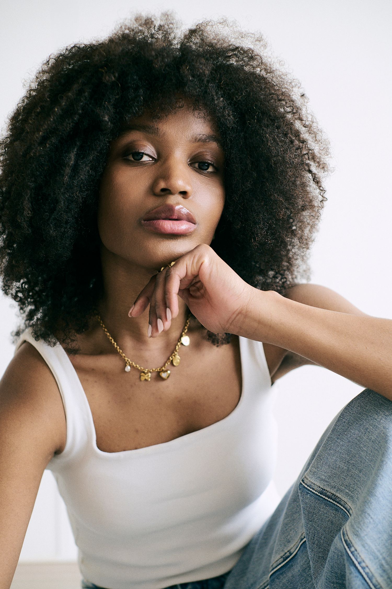 Young woman with natural afro hair wearing a white tank top and gold charm necklace, posing thoughtfully.