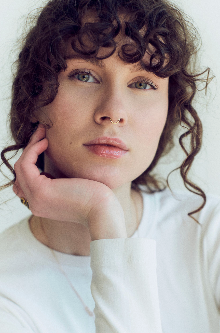 Young woman with curly brown hair and nose ring resting chin on hand, wearing white top against light background.