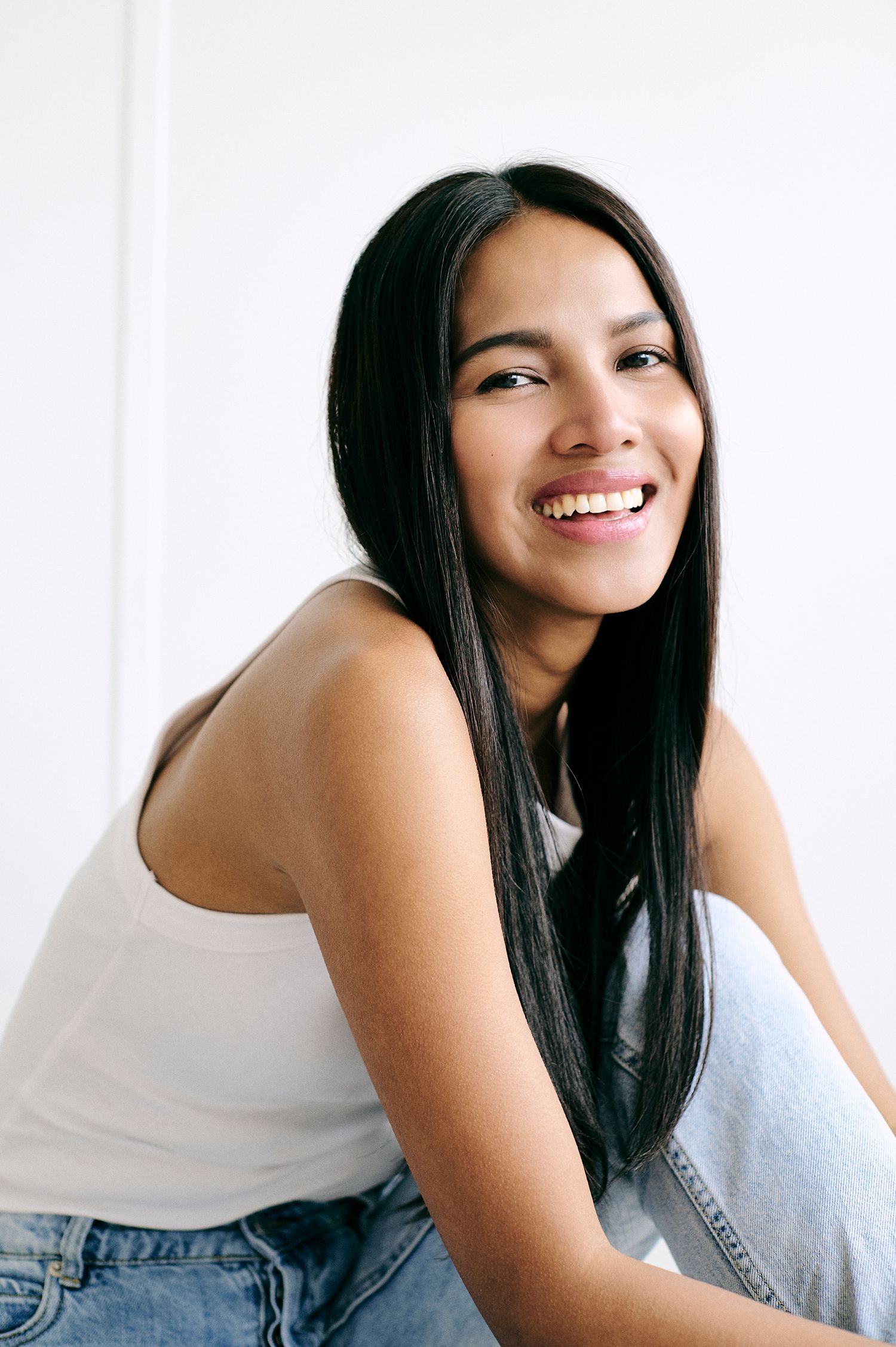 Smiling young woman with long black hair wearing a white tank top and jeans, sitting against a white background.