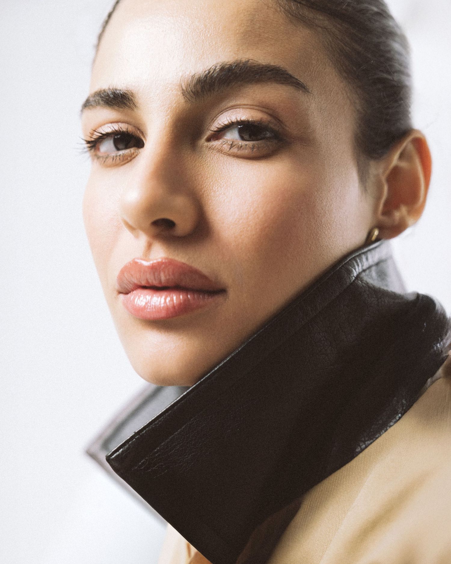 Close-up portrait of a young woman with glowing skin, subtle makeup, and a sleek updo, wearing a dark leather collar jacket.