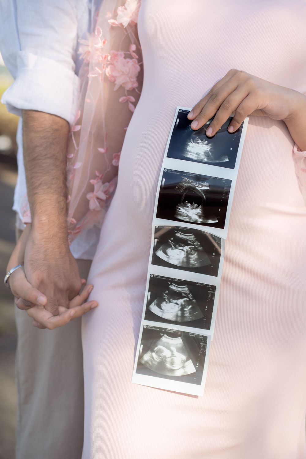 Pregnant couple holding hands outdoors, woman in pink dress displaying ultrasound sonogram photos against her baby bump.
