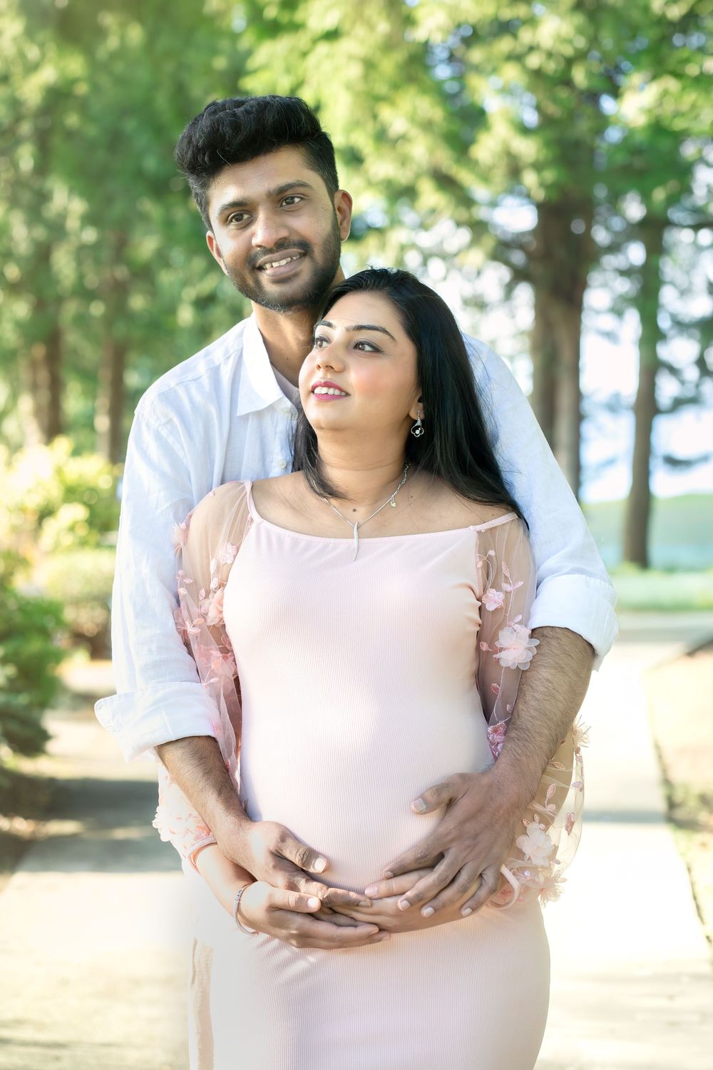 A happy couple in a park, man embracing pregnant woman in pink dress, both smiling outdoors.